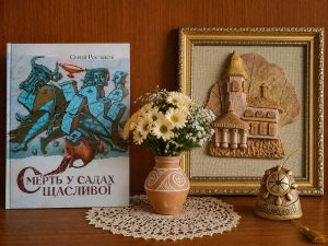 Книга Сергія Рослякова «Смерть у садах щасливої» на книжковій полиці поруч із декоративною композицією та букетом квітів